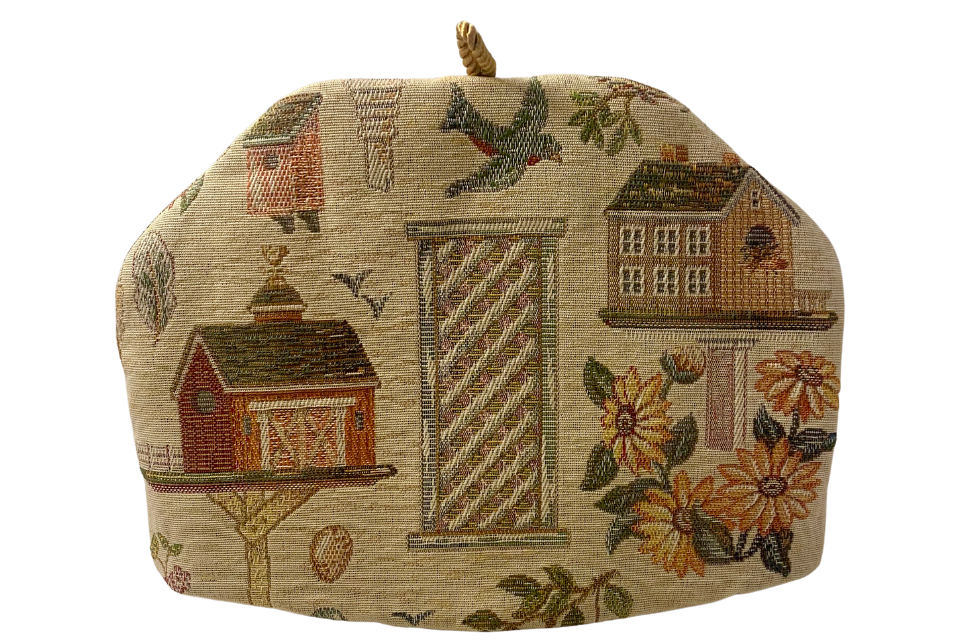 A beige fabric tea cozy featuring an embroidered bird house design with birds, flowers, and a rural house motif.