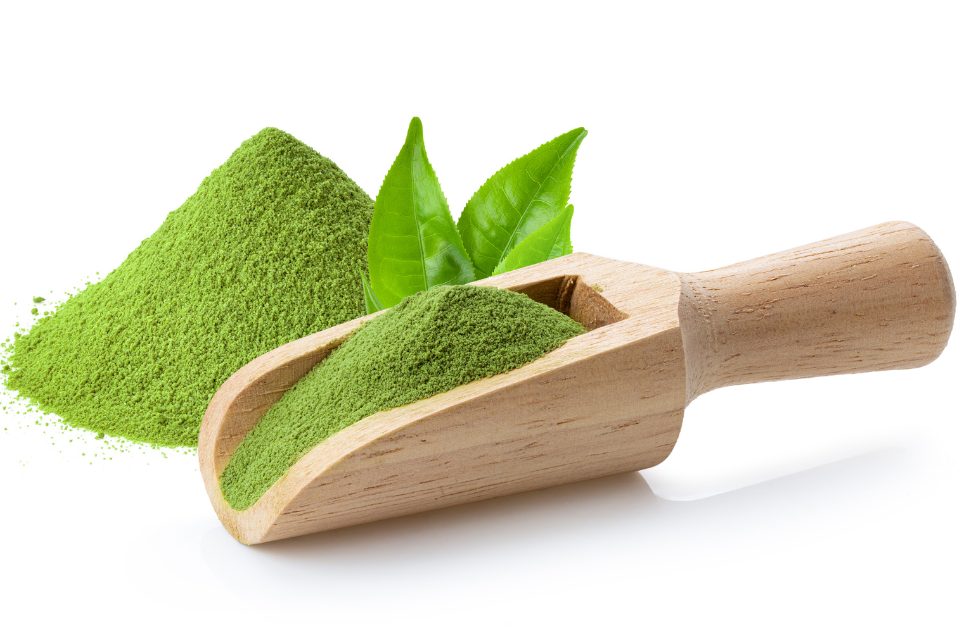 A wooden scoop filled with japanese green matcha powder, with fresh green tea leaves beside it.