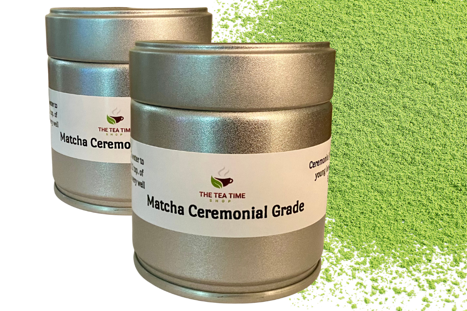 Two cylindrical metal canisters labeled 'Matcha Ceremonial Grade' from The Tea Time Shop, placed against a background with a matcha powder splash.