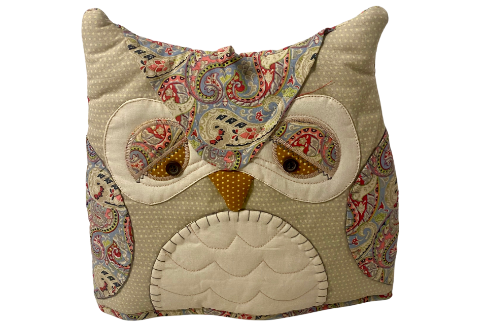 A fabric tea cozy with an owl design, featuring a multicolored pattern and a button eye detail.