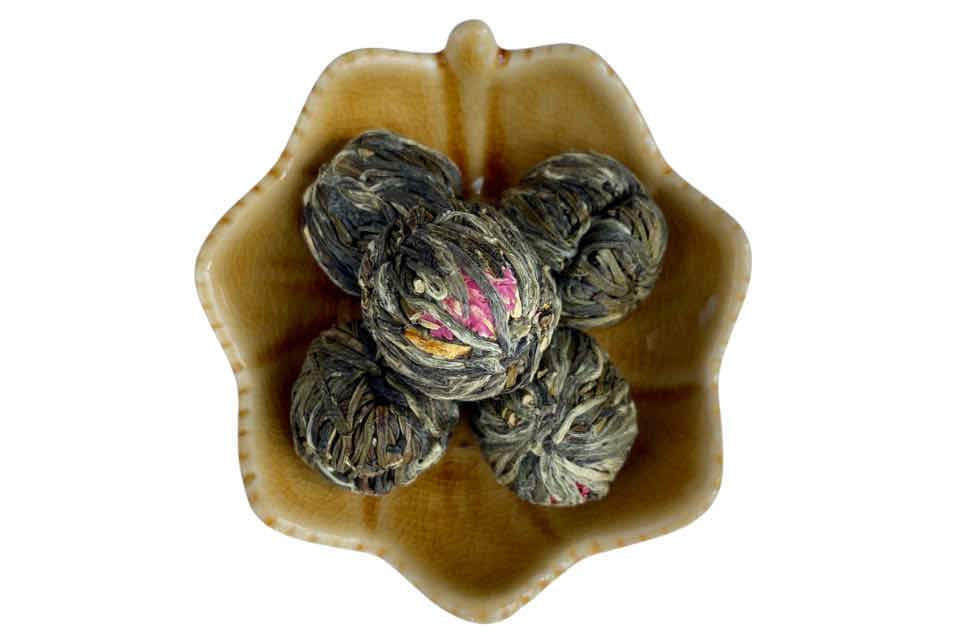 A collection of green tea balls with floral patterns, resembling opened tea leaves, arranged in a brown ceramic bowl.
